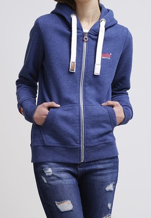 Vrouw draagt een blauwe hoodie met ritssluiting en witte koorden, gescheurde blauwe spijkerbroek, handen in de zakken van de hoodie, staand tegen een effen achtergrond.