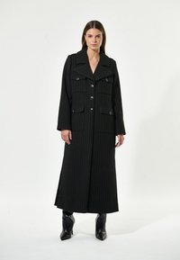 Cappotto lungo a righe nere con ampi revers, tasche frontali e chiusura con bottoni. Realizzato in tessuto texturizzato, presenta maniche lunghe e spacchi laterali.