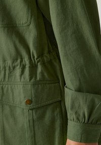 Lähikuva vihreästä takin hihasta ja taskusta, jossa on messinkinen nappilukko, näyttäen kankaan rakenteen ja ompeluyksityiskohdat.