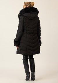 Frau trägt einen schwarzen Daunenmantel mit Fellbesatz an der Kapuze und den Manschetten, kombiniert mit schwarzen Leder-Stiefeln bis zum Knie, steht vor einem schlichten Hintergrund.