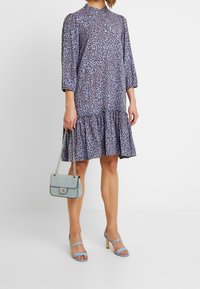 Abito blu con stampa floreale, orlo arricciato, maniche a tre quarti e colletto alto. Abbinato a una borsa a tracolla azzurra e tacchi a strappo.