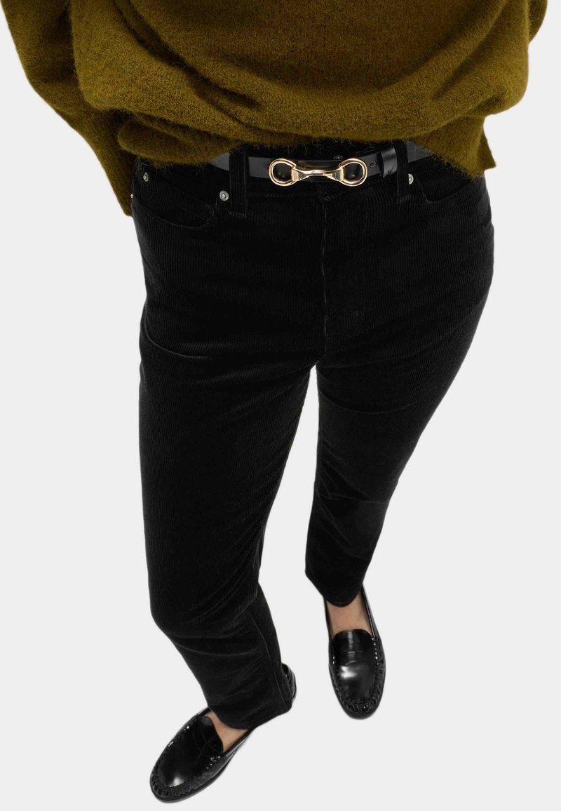 Schwarze Cordhosen mit schlankem Schnitt, ausgestattet mit Vordertaschen und einem schwarzen Gürtel mit einer goldenen Schnalle in Form einer Unendlichkeitsschleife. Schwarze Mokassins runden den Look ab.