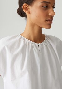 Weiße Bluse mit Rundhalsausschnitt, Raffung und zwei Knöpfen vorne. Hergestellt aus einem glatten, leichten Stoff mit kurzen Ärmeln.