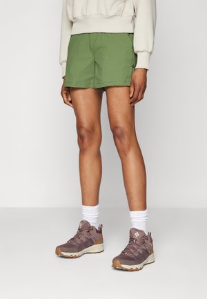 Persona con pantalones cortos verdes, calcetines blancos hasta el tobillo y zapatos de senderismo malva de pie frente a un fondo claro y liso.