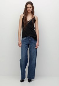 Musta pitsireunainen toppi yhdistettynä korkeavyötäröisiin, leveälahkeisiin sinisiin farkkuihin, joissa on vaalea pesu ja koristeltuja yksityiskohtia. Mustat kengät.