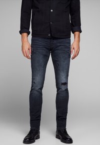Man i mörka tvättade skinny jeans med subtil slitage, svart knäppt jacka och svarta läder snörskor, som står mot en grå bakgrund.