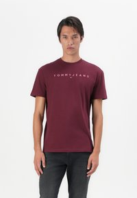 Bordeauxfarbene Baumwoll-T-Shirt mit kurzen Ärmeln, das das Logo "TOMMYJEANS" in Weiß zeigt. Rundhalsausschnitt, einfarbige Textur und reguläre Passform.