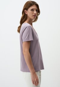T-shirt a maniche corte di colore lavanda realizzato in tessuto morbido. Presenta un colletto a V discreto e una vestibilità comoda, abbinato a pantaloni chiari.