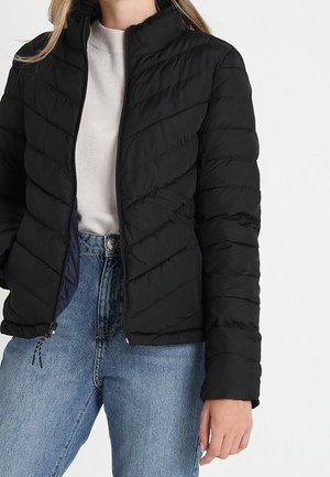 Kvinde iført en sort quiltet jakke med lynlås over en lys beige sweater og blå højtaljede jeans mod en ensfarvet hvid baggrund.
