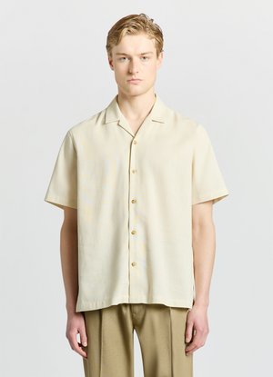 Jeune homme aux cheveux clairs portant une chemise beige à manches courtes boutonnée et un pantalon marron clair, debout devant un fond clair uni.