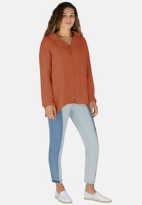Junge Frau steht mit einem rostorangefarbenen Langarmshirt, zweifarbigen blauen Skinny-Jeans, weißen Slippern und goldener Schmuck.