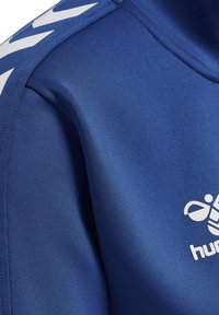 Blauwe sportkledingstof met een gladde textuur, featuring een groot wit logo en een contrasterend wit chevronpatroon langs de schoudernaad.
