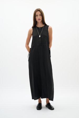 Mladá žena stojí a má na sebe čierne maxi šaty bez rukávov, čierne slip-on topánky a príveskový náhrdelník na jednoliatom bielom pozadí.