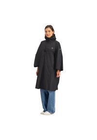 Zwarte waterdichte poncho met een capuchon, voorvak en verstelbare zijknoppen, gedragen over blauwe wijde jeans en witte schoenen.