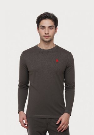 Junger Mann mit dunklem Haar, der ein enges, dunkelgraues Langarmshirt mit einem kleinen roten Logo auf der Brust trägt, steht vor einem schlichten Hintergrund.