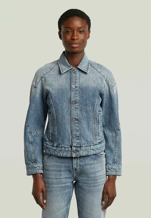 Frau mit kurzen Haaren, die eine hellblaue, geknöpfte Jeansjacke und passende Jeans trägt und vor einem einfarbigen hellen Hintergrund steht.