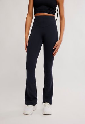 Mujer con leggins negros acampanados, top corto negro y zapatillas blancas, de pie frente a un fondo liso.
