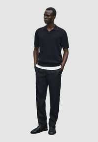 Polo shirt nero a coste con colletto marrone, abbinato a pantaloni scuri e sandali neri. Il modello è in piedi con le mani in tasca contro uno sfondo chiaro.