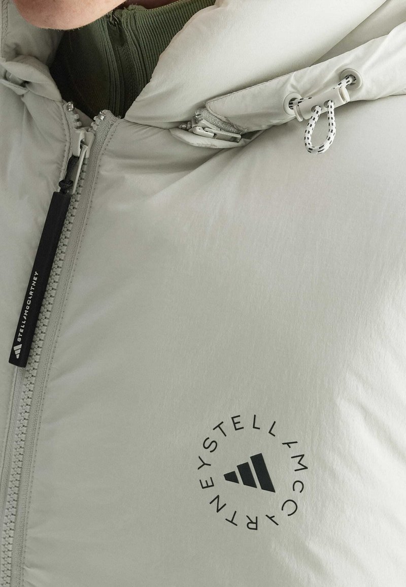 Chaqueta puffy de color gris claro con cremallera, cuenta con capucha, cordones ajustables y un logotipo negro circular en la zona del pecho.