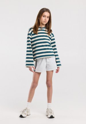 Jong persoon staand, gekleed in een groen-wit gestreepte lange mouw top, lichtgrijze korte broek met zwarte strepen, witte sokken en sneakers.