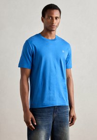 T-shirt azul de manga curta feito de algodão macio, com decote redondo e um pequeno logotipo branco no peito esquerdo. Usado com jeans de ganga desbotados.