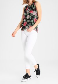 Haut sans manches floral en noir avec des motifs roses et verts, associé à un jean skinny blanc et des baskets noires avec des semelles blanches.