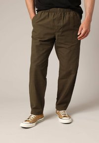Homme portant un pantalon décontracté vert olive et des baskets en toile beige avec des lacets blancs, se tenant avec une main dans la poche.