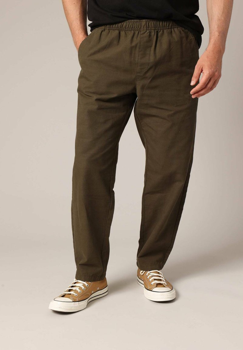 Homme portant un pantalon décontracté vert olive et des baskets en toile beige avec des lacets blancs, se tenant avec une main dans la poche.