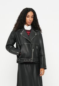 Μαύρο δερμάτινο μπουφάν μηχανής με κομμένο σχέδιο, υπερμεγέθη παλτό και μπροστινό φερμουάρ. Διαθέτει πλαϊνές τσέπες και ζώνη στη μέση.