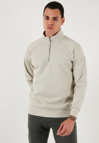 Svijetlo sivi flis pulover s visokim ovratnikom i zatvaranjem na pola munje. Ima raglan rukave i suptilan logo na prsima. Glatka tekstura.