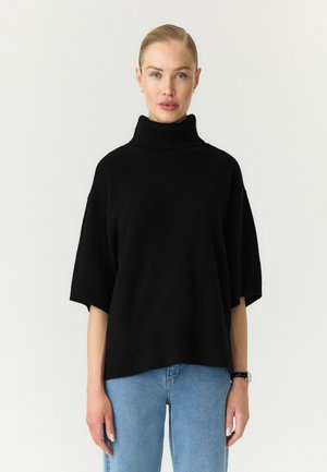 Femme blonde aux cheveux tirés en arrière portant un pull à col roulé noir à manches courtes et un jean bleu clair, debout devant un fond uni.