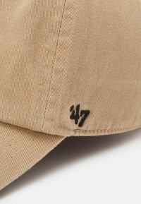 Beige cap laget av teksturert stoff, med fremtredende sømmer og et svart brodert "47" på siden. Buet skygge, uformell design.