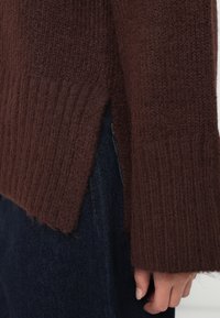 Καφέ πλεκτό πουλόβερ με ραβδώσεις, με πλευρικές σχισμές και μακριά μανίκια. Φορεμένο πάνω από σκούρα τζην, αναδεικνύοντας τη μαλακή υφή και το χαλαρό στυλ.