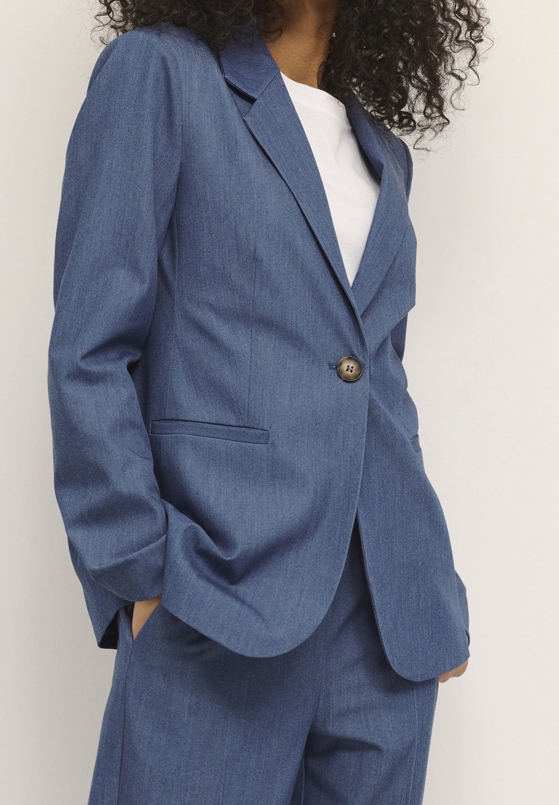 Personne portant un blazer bleu à un bouton et un pantalon assorti, les mains dans les poches sur une chemise blanche, sur un fond uni.