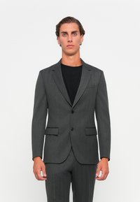 Donkergrijze visgraatcolbert met notch-revers, twee voorzakken en een enkele knoopsluiting, gecombineerd met een zwart shirt.
