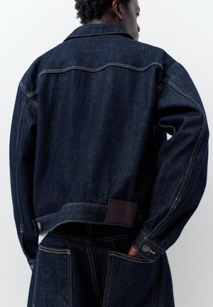 Moški, oblečen v temno moder denim jopič in ujemajoče kavbojke, stoji z eno roko v zadnjem žepu, obrnjen stran od kamere.