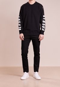 Sweat-shirt noir avec texte blanc "VERSUS" sur les manches, associé à un pantalon noir et des baskets blanches. Tissu lisse, col rond.