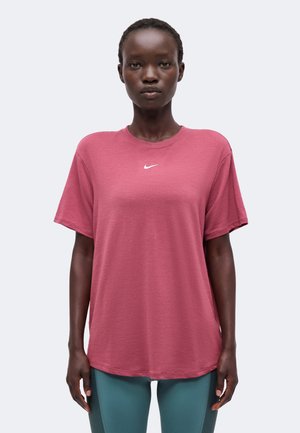 Tmavá osoba stojaca čelom vpred, oblečená v voľné ružové tričko Nike a tyrkysové športové legíny na jednoduchom pozadí.