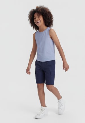 Glimlachend kind met krullend haar aan het wandelen, gekleed in een lichtblauw mouwloos shirt, marineblauwe cargoshorts en witte hoge sneakers.