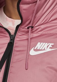 Rosa vadderad jacka med dragkedja och justerbar huva. Inkluderar en vit Nike-logga på bröstet och en texturerad tygyta.