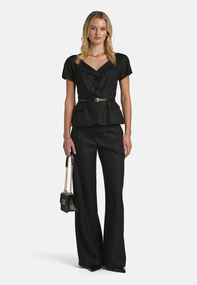 Femme portant un haut noir à manches courtes boutonné avec ceinture, pantalon noir à jambes larges, sac à main noir avec chaîne dorée, debout devant un fond blanc.