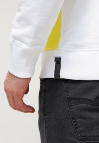 Sudadera blanca con un acento amarillo en el lateral; puños y dobladillo acanalados; presenta un botón decorativo negro en la parte inferior. Jeans oscuros son visibles.