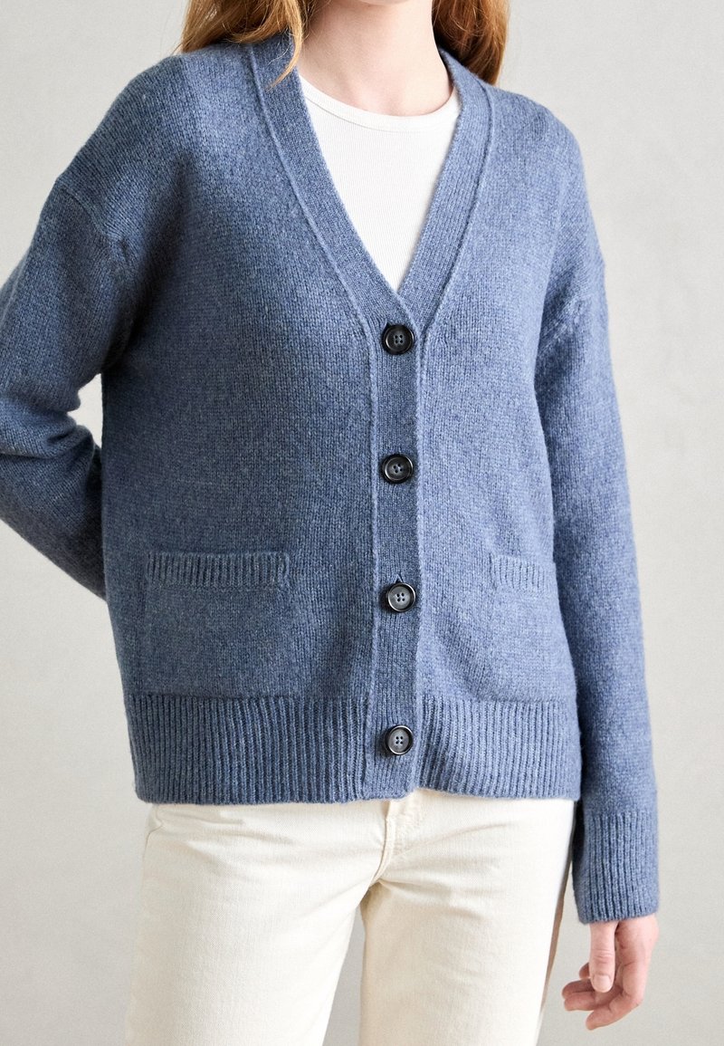 Femme portant un cardigan en maille bleu boutonné sur une chemise blanche et un pantalon crème, debout devant un fond clair uni.