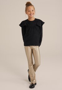 Črn strukturiran pulover z nabranimi rokavi, v kombinaciji s prilegajočimi zlatimi usnjenimi hlačami in črnimi masivnimi čevlji. Enostavna ozadja.