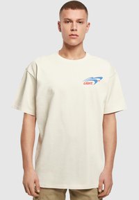 Creme oversized T-shirt, lavet af bomuldsmateriale, rund hals, korte ærmer, med et blåt og rødt grafisk logo på venstre brystområde.