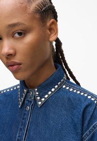 Camicia in denim di colore blu scuro, con borchie metalliche lungo il colletto e un design classico con bottoni. Tessuto strutturato con una vestibilità definita.