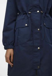 Marineblauwe jas met een taillerende taille, elastische manchetten, meerdere voorzakken en goudkleurige drukknopen langs de sluiting en zakken.