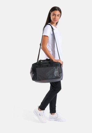 Donna che indossa una t-shirt bianca, pantaloni neri e sneakers bianche con una borsa duffel nera Arena sulla spalla.