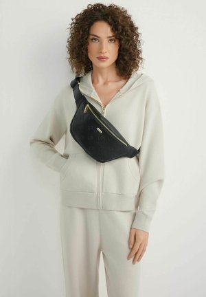 Kobieta o kręconych włosach, ubrana w beżową bluzę z kapturem i spodnie, niesie czarną torbę na ramię z złotymi zamkami, przewieszoną przez klatkę piersiową.