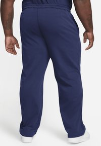 Námořnické modré tepláky vyrobené z měkkého materiálu, s volným střihem, elastickým pasem a bočními kapsami. Bílé tenisky viditelné dole.